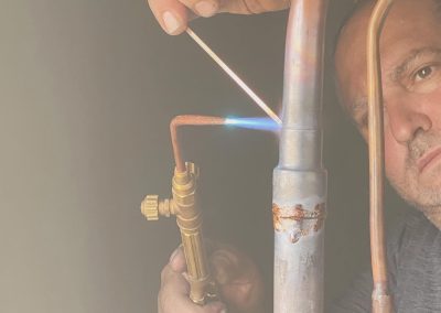 A man welds copper pipes using a torch and welding rod, concentrating on his work.