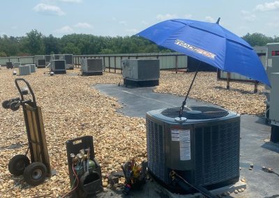 An air conditioning unit on a gravel rooftop is shaded by a blue TraneCare umbrella, with repair tools nearby.
