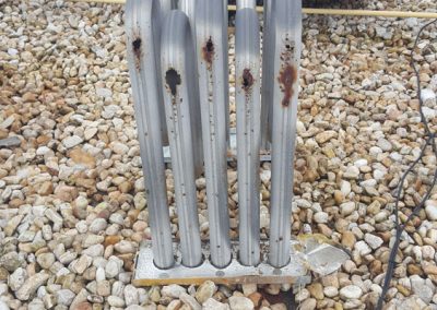 Metal pipes with rust spots stand upright on a rocky rooftop, next to cables and HVAC equipment.