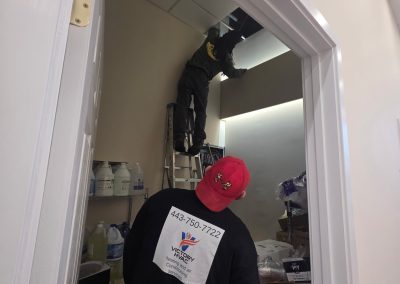 Two workers in a storage room; one on a ladder fixing the ceiling, another standing below watching.