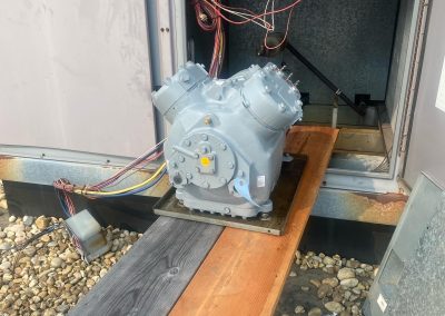 Large metal compressor on wooden planks outside electrical cabinet with exposed wiring and rocks below.