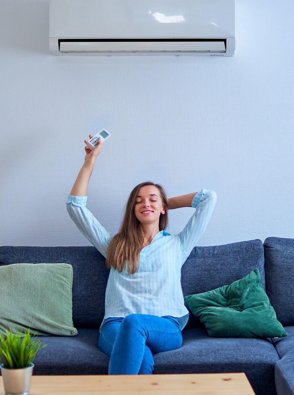 Woman relaxing on a sofa, holding up a remote, sitting under an air conditioner on a white wall.