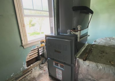 A gray furnace unit stands by a damaged wall and window in a room with exposed pipes and wooden floor.
