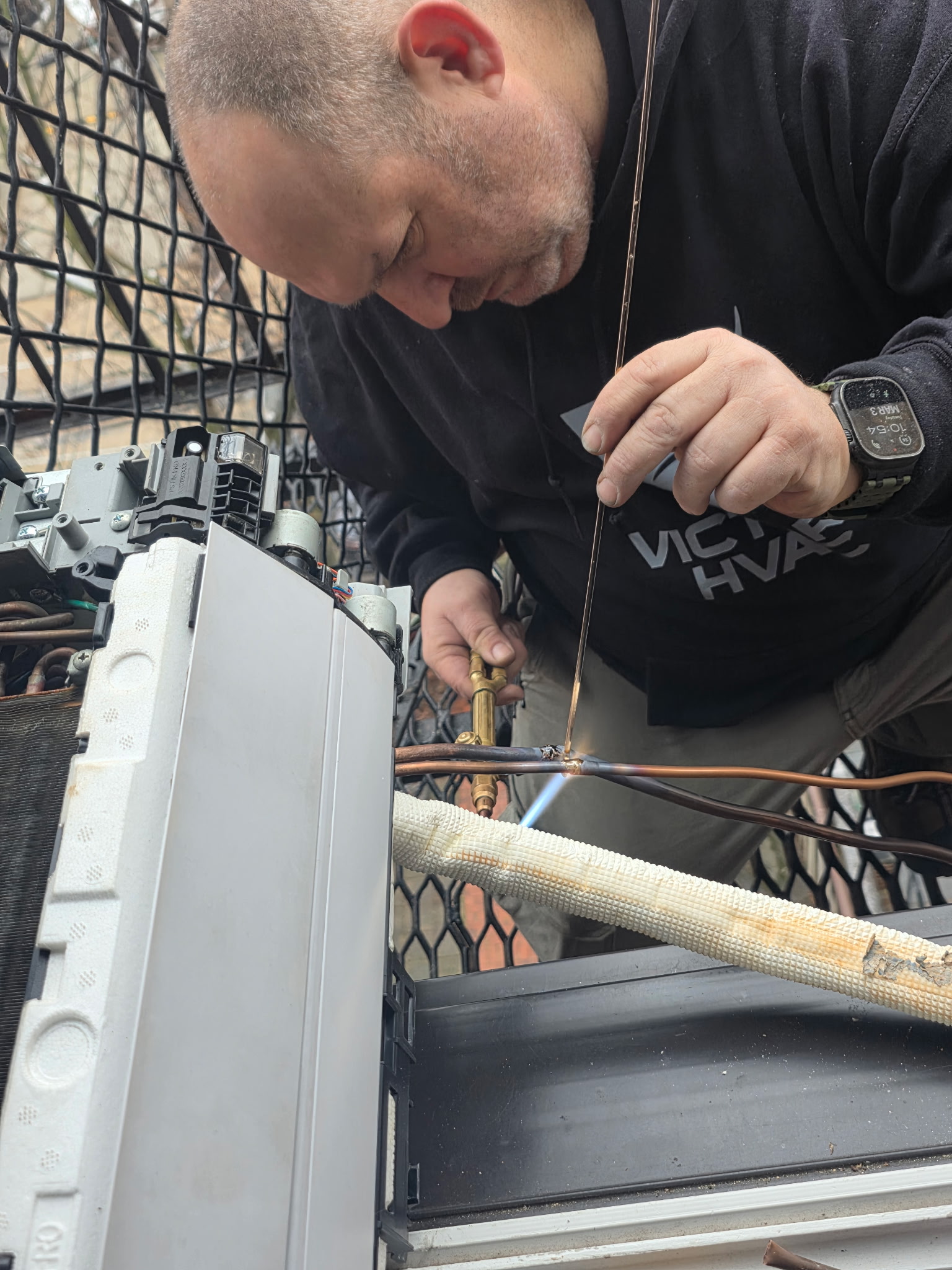 A man uses a torch to solder pipes on an HVAC unit, wearing a black sweatshirt and a smartwatch.
