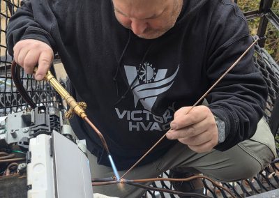 A man in a hoodie solders copper pipes on an outdoor HVAC unit using a blowtorch.