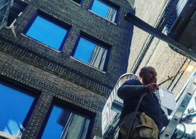 Man working on a ladder beside a tall brick building, holding equipment with blue sky reflected in windows.