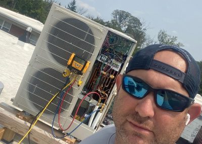 Man in sunglasses and cap working on an outdoor air conditioning unit on a rooftop under a sunny sky.