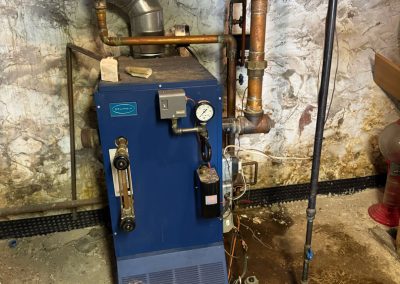 A blue boiler with pipes, gauges, and wires in a basement with a stone wall and stained floor.