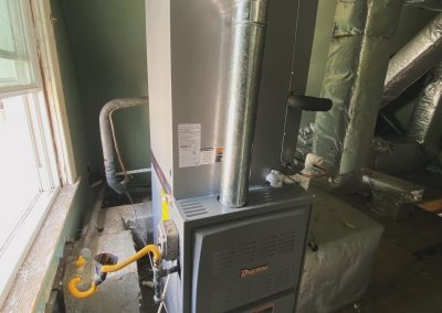 A gas furnace with metal ductwork installed in an unfinished room with green walls and exposed insulation.
