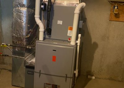 A gray furnace unit with white pipes installed in a basement with concrete walls and floor.