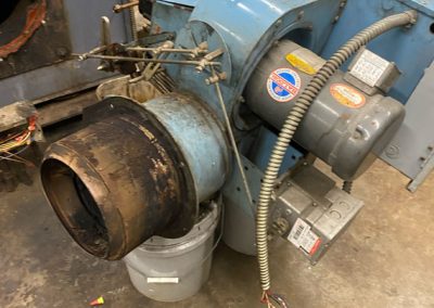 Industrial blower motor assembly with wires and gauges, placed on a workshop floor beside a metal bucket.