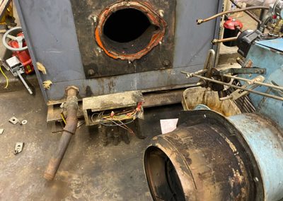 Disassembled industrial burner and boiler with visible open port and removed burner on a workshop floor.