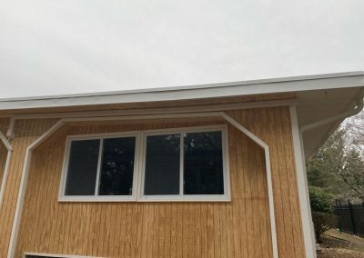 A wooden house exterior with three windows and an overhanging white roof under a cloudy sky.