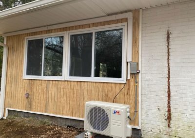 A tan wooden house wall with a large window and an outdoor AC unit installed below it.