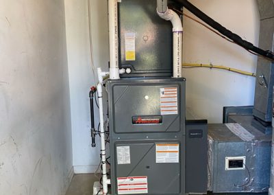 A gray HVAC furnace unit stands in a utility room with pipes and ductwork attached.