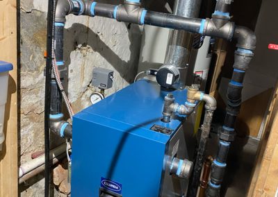 Blue boiler system with metal pipes and valves in a basement with stone walls and unfinished wooden framing.