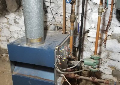 Old furnace with exposed wiring and pipes in a basement with a stone wall and dirt floor.
