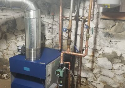 A blue furnace with copper pipes in a basement with stone walls and exposed ductwork.