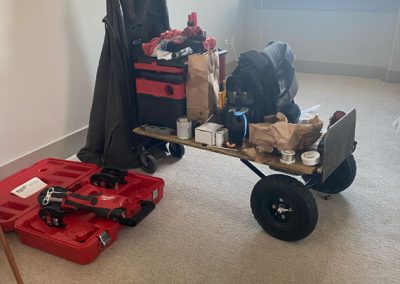 A utility cart with tools, bags, and boxes next to a window in a carpeted room.