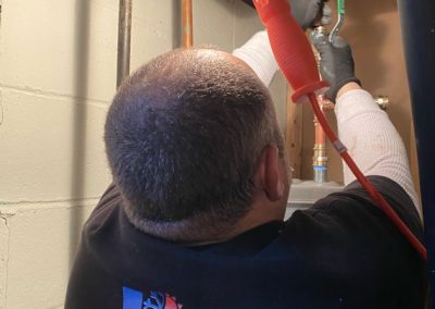 A plumber in gloves works on pipes near a ceiling, wearing a Victory HVAC shirt and using a red tool.