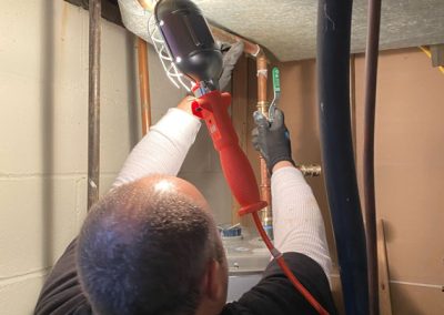 Person using a work light and wrench to fix copper pipes near a ceiling in a utility room.