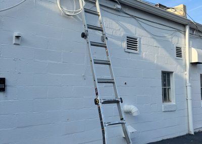 A tall ladder leans against a white brick building with cables and vents visible on the wall.