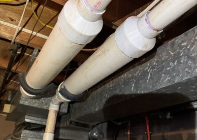 Two white PVC pipes run parallel along a basement ceiling, connected to ductwork and surrounded by wires.