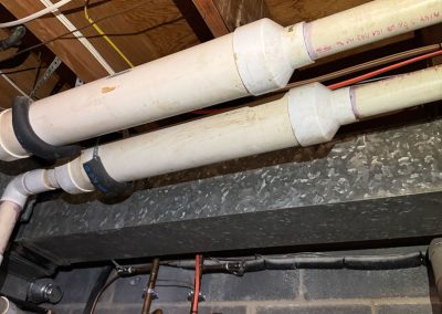 White PVC pipes run along an unfinished basement ceiling with exposed wood beams, wires, and metal ductwork.