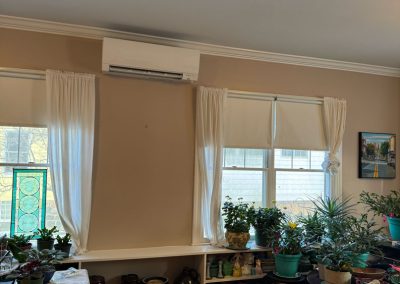 Bright room with potted plants on shelves and tables in front of windows with white curtains and blinds.