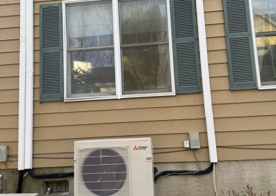 A heat pump unit is mounted on a stand outside a house with tan siding and green shutters.