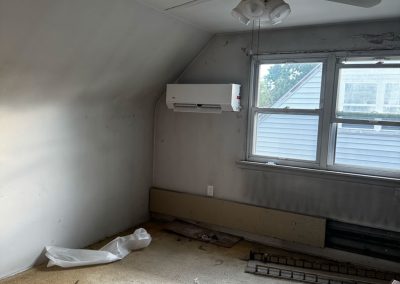 Small, empty room with stained carpet, ceiling fan, wall AC, and boxes near a window with peeling paint.
