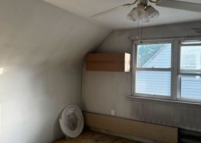 An empty, dusty room with stained carpet, a ceiling fan, window, and a cardboard box on the wall.