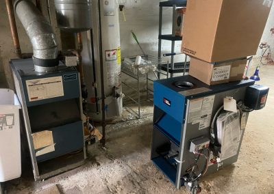 Two furnaces and a water heater in a basement; one furnace is partially assembled with a box on top.