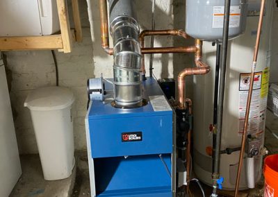 A blue home boiler with metal pipes in a basement, next to a water heater and a white trash bin.