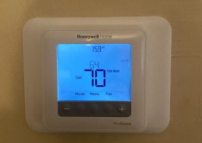 A lit Honeywell Home thermostat displays a temperature of 70°F and the time 1:59 PM on a beige wall.