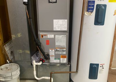 A water heater and furnace installed side by side in a basement utility area with visible pipes and labels.