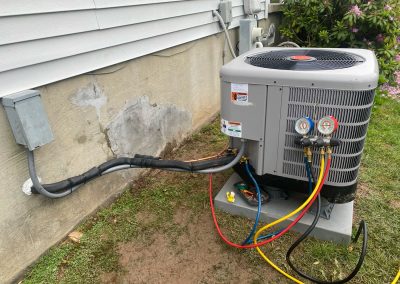 Outdoor air conditioning unit with gauges attached, next to a house exterior and electrical box.