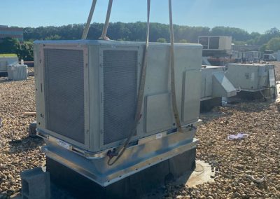 A large HVAC unit being lifted onto a gravel rooftop with straps under a clear sky.