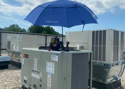 A blue umbrella shades tools on top of an outdoor HVAC unit, surrounded by rocks and other units.
