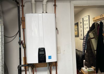 A white tankless water heater with pipes is mounted on a wall in a utility room next to an open doorway.