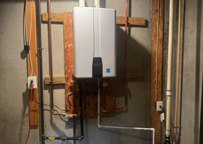A white tankless water heater is mounted on a basement wall with pipes connected and a sump pump nearby.