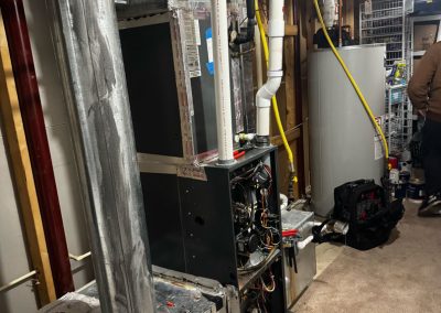 A furnace with its panel open for maintenance in a basement, near a water heater and tools.