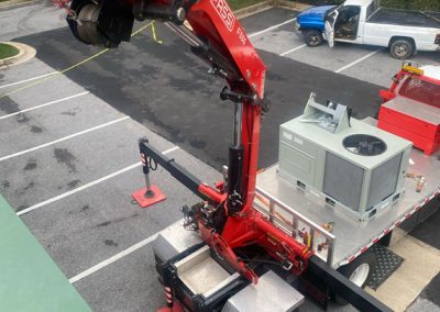 A red crane lifts HVAC equipment onto a building from a flatbed truck in a parking lot.