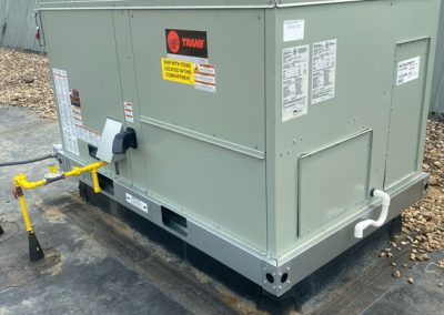 Large gray Trane HVAC unit on a rooftop, surrounded by gravel and a sloped metal wall in the background.