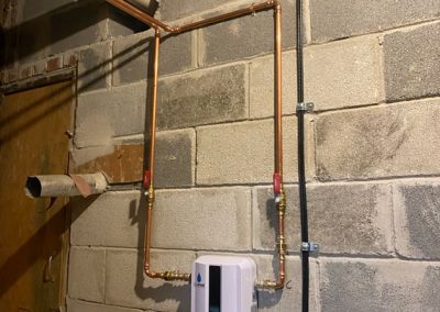 Small tankless water heater with copper pipes mounted on an unfinished cinder block basement wall.