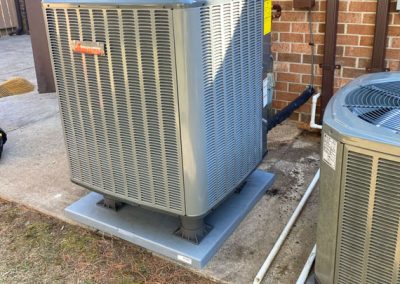 Gray outdoor air conditioning unit installed next to a brick building, with pipes and another unit nearby.