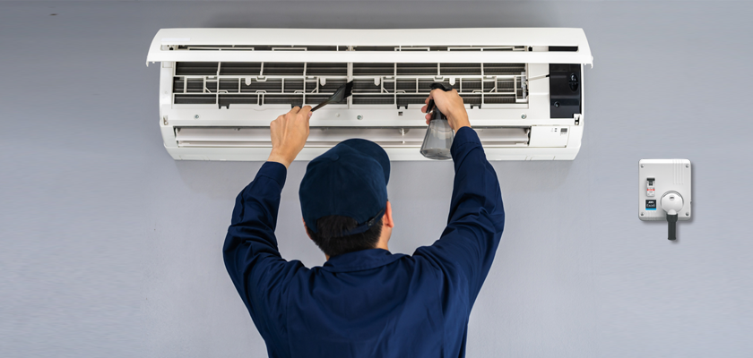 A technician in blue uniform cleans an air conditioner unit mounted on a wall with a brush and spray bottle.