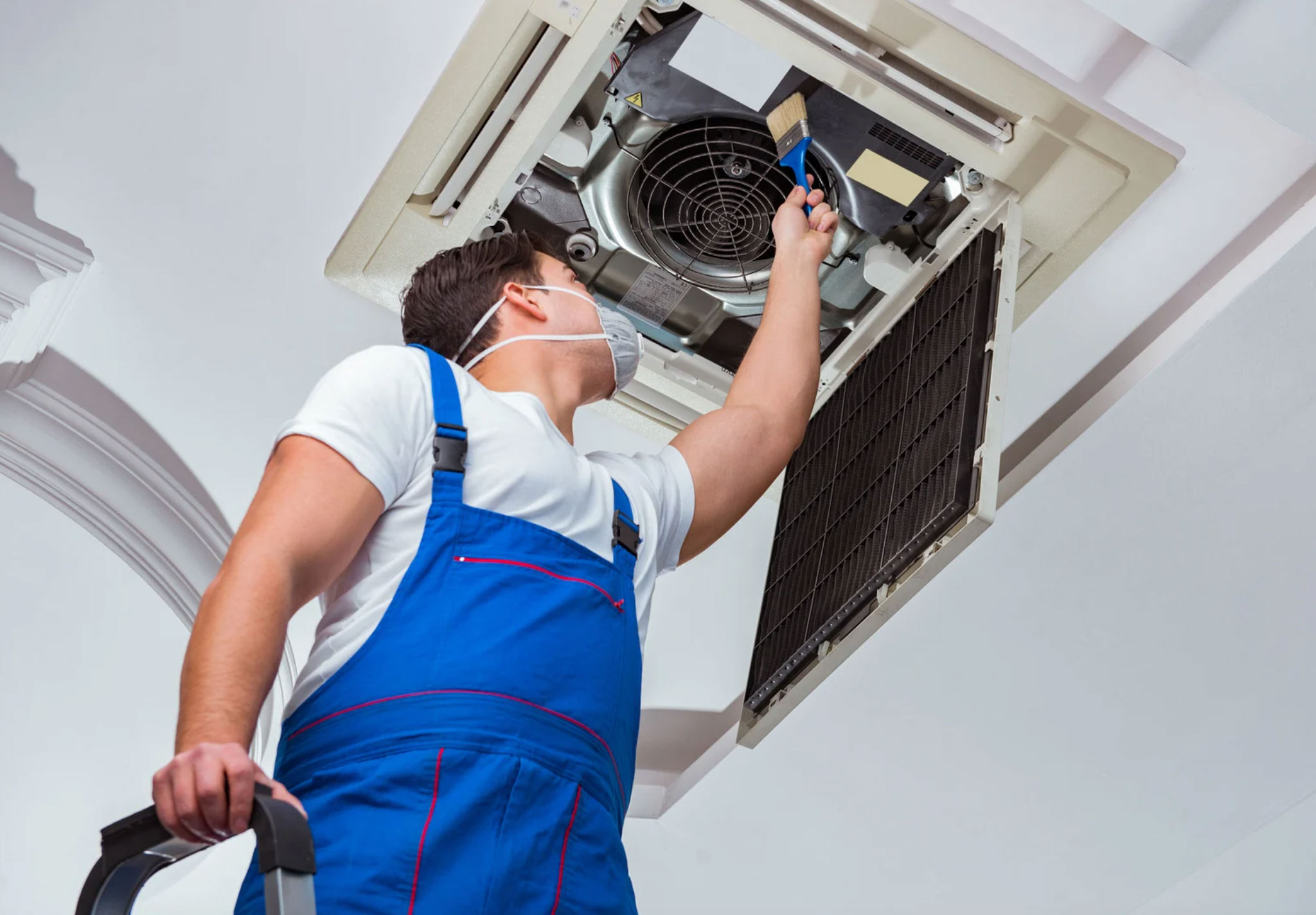 A technician in blue overalls cleans an air conditioner on the ceiling while wearing a face mask.