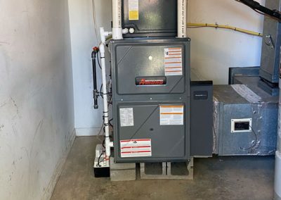 Gray Amana furnace installed in a utility room, sitting on concrete blocks with visible labels and pipes.