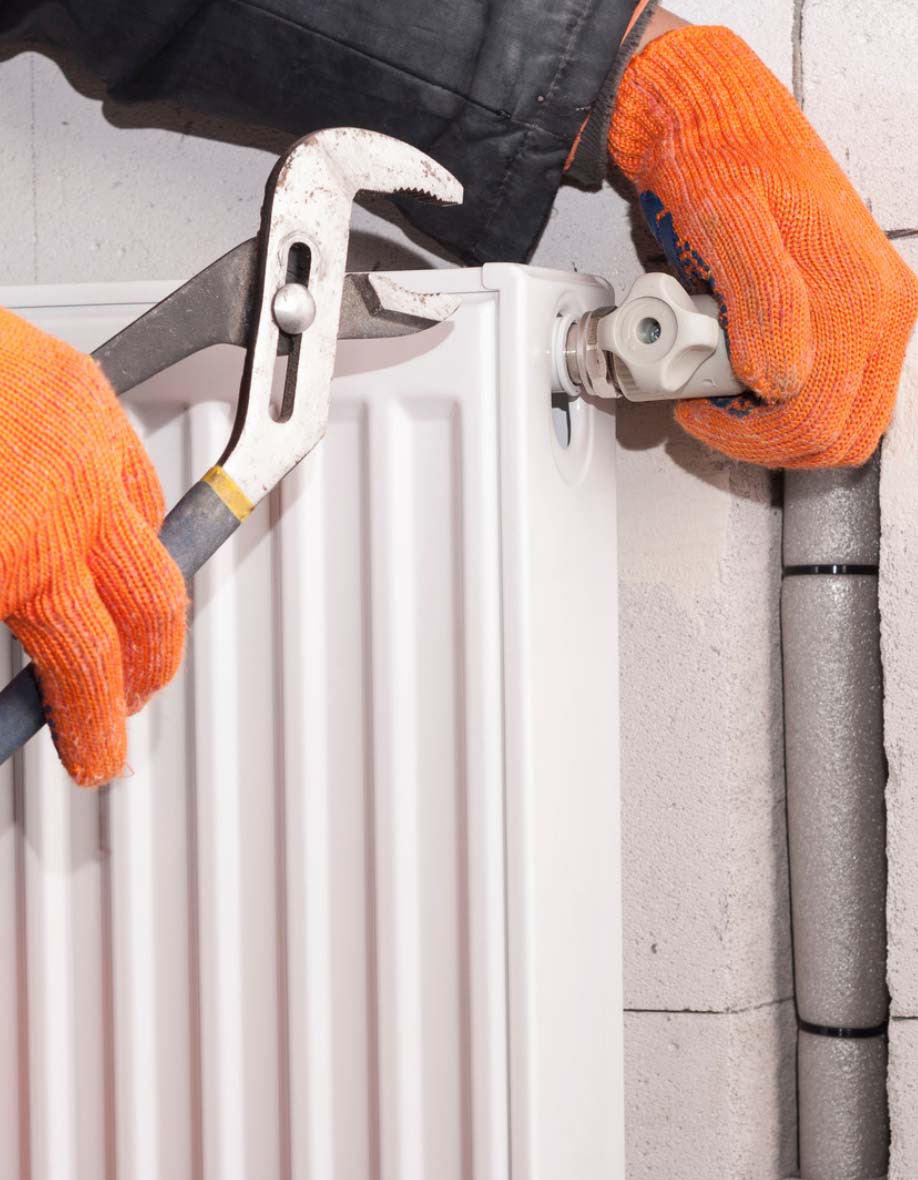 Person in orange gloves using a wrench to tighten a valve on a white radiator near a wall and metal pipe.